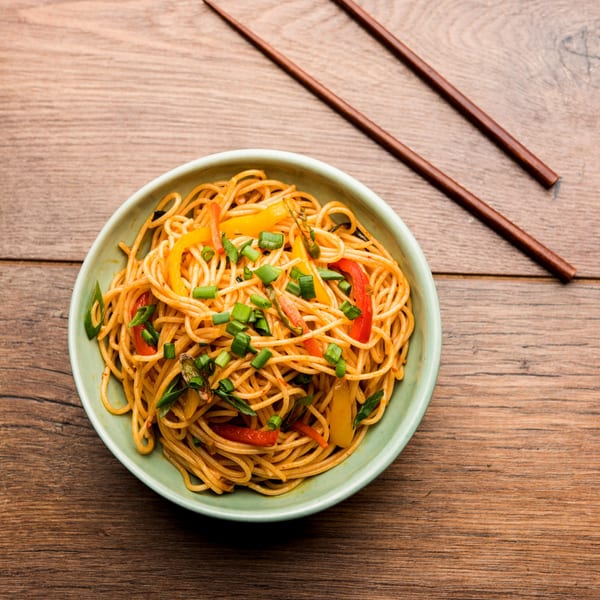 Szechuan noodles with bell peppers and green onions in a light green bowl, with chopsticks on a wooden table.