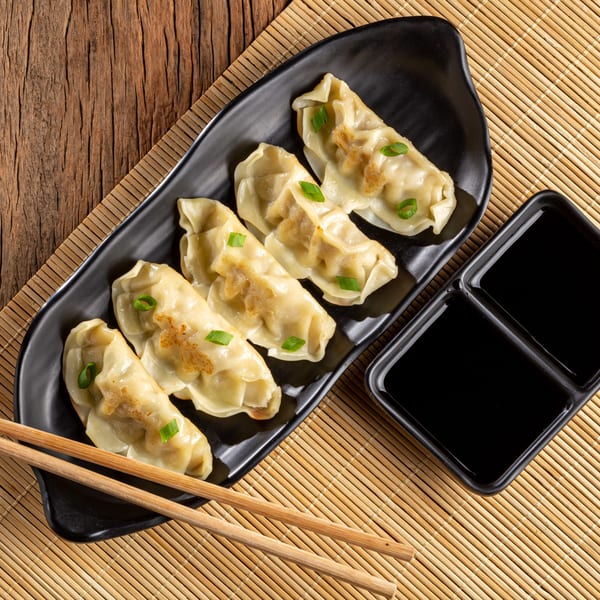Pan-fried Szechuan dumplings with green onion garnish, soy sauce, and chopsticks on a bamboo mat.